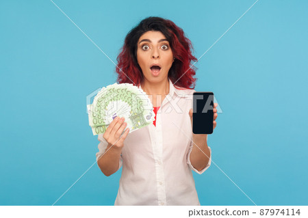 Online payment. Astonished surprised woman with fancy red hair holding cell phone and euro banknotes, shocked by money cashback and mobile application. indoor studio shot isolated on blue background 87974114
