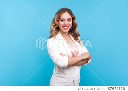 Portrait of joyful bright businesswoman with wavy hair in white jacket smiling at camera, looking fashionable confident and successful with crossed hands. indoor studio shot isolated, blue background 87974130