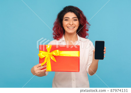 Gift order online. Portrait of happy smiling woman with fancy red hair showing wrapped box and cellphone, looking satisfied with present, mobile service. indoor studio shot isolated on blue background 87974131