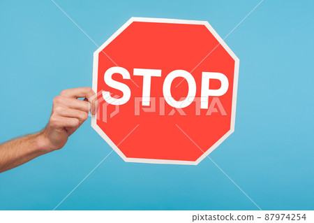 Closeup of male hand holding Stop symbol, showing red traffic sign, warning about problems, concept of ban, denial, prohibited to go, forbidden way. indoor studio shot isolated on blue background Closeup of male hand holding Stop symbol, showing red traffic sign, warning about problems, concept of ban, denial, prohibited to go, forbidden way. indoor studio shot isolated on blue background 87974254