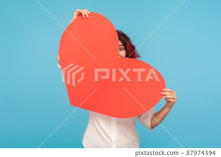 Love you on Valentine day. Young playful unknown woman hiding behind large heart and looking with funny curious eye, holding symbol of romantic relation. indoor studio shot isolated on blue background Love you on Valentine day. Young playful unknown woman hiding behind large heart and looking with funny curious eye, holding symbol of romantic relation. indoor studio shot isolated on blue background 87974394