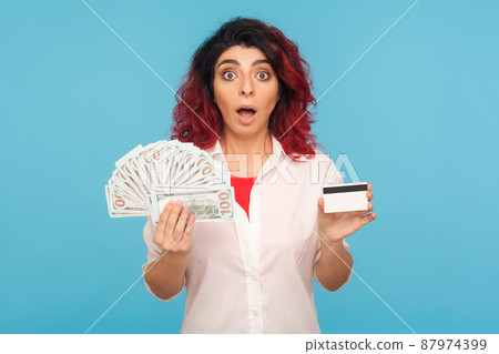 Wow, bank loan for shopping. Portrait of surprised hipster woman with fancy red hair holding credit card, dollar bills and looking with amazed shocked face. studio shot isolated on blue background Wow, bank loan for shopping. Portrait of surprised hipster woman with fancy red hair holding credit card, dollar bills and looking with amazed shocked face. studio shot isolated on blue background 87974399