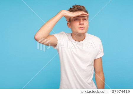 Far away vision. Portrait of inquisitive man in casual white t-shirt peering long distance and holding hand over his eyes, watching with attentive view. indoor studio shot isolated on blue background Far away vision. Portrait of inquisitive man in casual white t-shirt peering long distance and holding hand over his eyes, watching with attentive view. indoor studio shot isolated on blue background 87974419