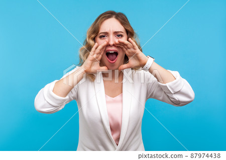 Attention please, Portrait of beautiful woman with wavy hair in white jacket holding hands near wide open mouth and shouting, loudly screaming announcement. studio shot isolated on blue background 87974438