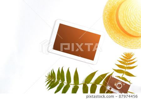 Straw hat with green leaves, tablet and old camera on white background, Summer background. Top view. Sun flare Straw hat with green leaves, tablet and old camera on white background, Summer background. Top view. Sun flare 87974456