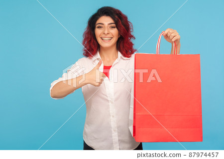 Great shopping. Portrait of excited shopaholic hipster woman with fancy red hair holding packages and showing thumbs up, satisfied with weekend sale. indoor studio shot isolated on blue background 87974457