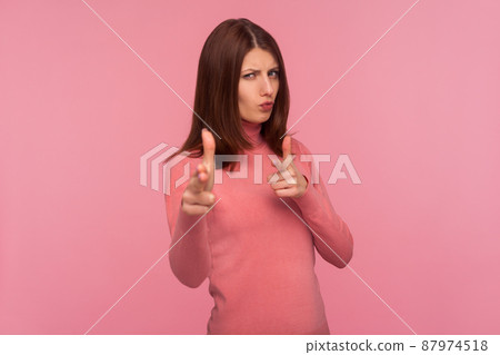 Assertive self confident woman with brown hair in pink sweater showing guns with her fingers seriously looking at camera aiming you, self defence. Indoor studio shot isolated on pink background 87974518