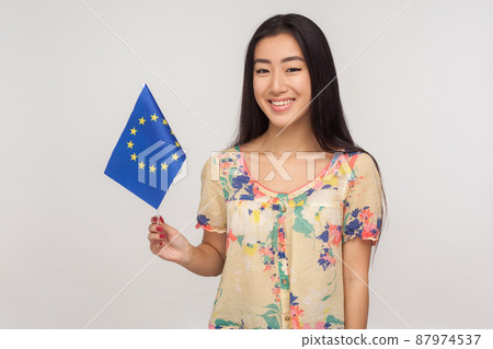 EU association. Happy asian tourist girl with brunette hair in summer blouse holding flag of European Union, immigration and travelling in Europe. indoor studio shot isolated on gray background 87974537