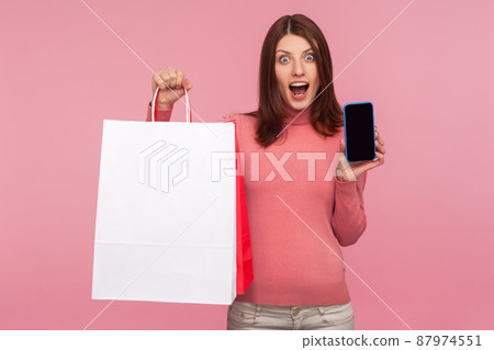 Shocked surprised woman holding paper bags and smartphone in hands, looking at camera with excitement, mobile shopping. Indoor studio shot isolated on pink background 87974551
