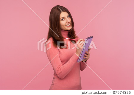 Positive happy woman making to do list writing down ideas on notepad in her hand looking at camera with toothy smile. Indoor studio shot isolated on pink background Positive happy woman making to do list writing down ideas on notepad in her hand looking at camera with toothy smile. Indoor studio shot isolated on pink background 87974556