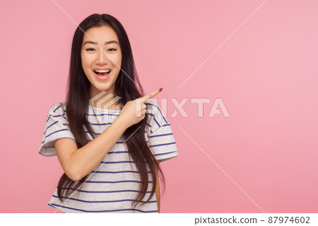Look at advertisement, Portrait of charming asian girl in striped t-shirt pointing to side and smiling, showing copy space, empty place for commercial. indoor studio shoot isolated on pink background 87974602