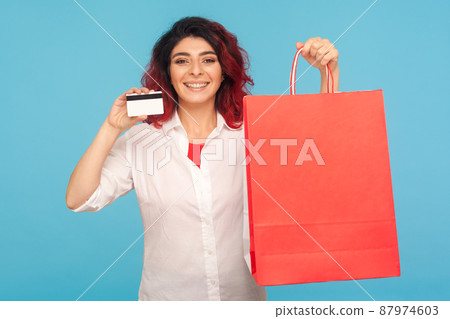 Bank loan for purchase. Portrait of joyful shopaholic hipster woman with fancy red hair holding shopping bags and credit card, satisfied with cashback. indoor studio shot isolated on blue background 87974603