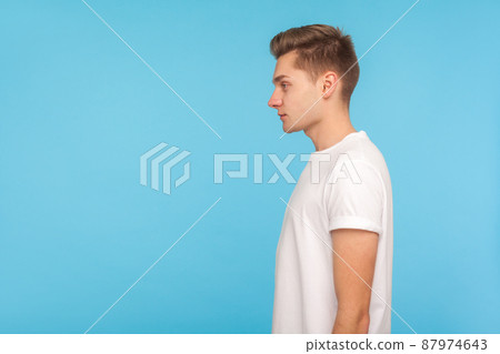 Profile of young serious guy with stylish hairdo in casual white t-shirt standing, looking to the side with attentive focused unsmiling expression. indoor studio shot isolated on blue background Profile of young serious guy with stylish hairdo in casual white t-shirt standing, looking to the side with attentive focused unsmiling expression. indoor studio shot isolated on blue background 87974643