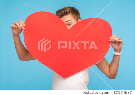 Romantic congratulation on Valentine day. Young man peeking out of big red paper heart, flirting, looking playfully to camera, declaration of love. indoor studio shot isolated on blue background 87974687
