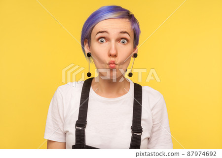 Portrait of amusing hipster girl with violet short hair in denim overalls making fish face with goofy comical expression, silly grimace, having fun. indoor isolated on yellow background, studio shot Portrait of amusing hipster girl with violet short hair in denim overalls making fish face with goofy comical expression, silly grimace, having fun. indoor isolated on yellow background, studio shot 87974692