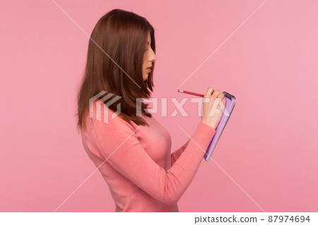 Profile portrait serious focused woman with brown hair making notes with pencil in notebook, writing down thoughts, making to do list. Indoor studio shot isolated on pink background Profile portrait serious focused woman with brown hair making notes with pencil in notebook, writing down thoughts, making to do list. Indoor studio shot isolated on pink background 87974694