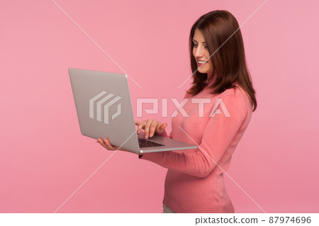 Smiling positive woman with brown hair in pink sweater working on laptop, freelance job, online studying. Indoor studio shot isolated on pink background Smiling positive woman with brown hair in pink sweater working on laptop, freelance job, online studying. Indoor studio shot isolated on pink background 87974696