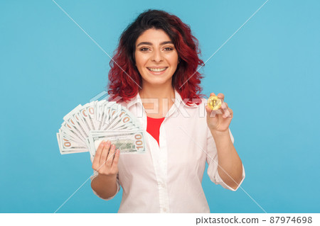Cryptocurrency exchange. Joyful smiling happy woman with fancy red hair holding bitcoin and dollar banknotes, digital money, electronic commerce. indoor studio shot isolated on blue background 87974698