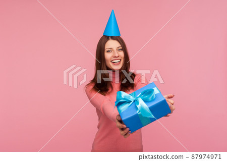 Happy brunette woman in pink sweater with party cone on head giving gift box to camera and smiling, congratulating on holiday. Indoor studio shot isolated on pink background 87974971