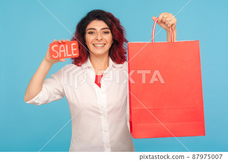 Discount in fashion store. Nice cheerful hipster woman with fancy red hair showing sale inscription and shopping bags, enjoying black friday, low prices. indoor studio shot isolated on blue background Discount in fashion store. Nice cheerful hipster woman with fancy red hair showing sale inscription and shopping bags, enjoying black friday, low prices. indoor studio shot isolated on blue background 87975007