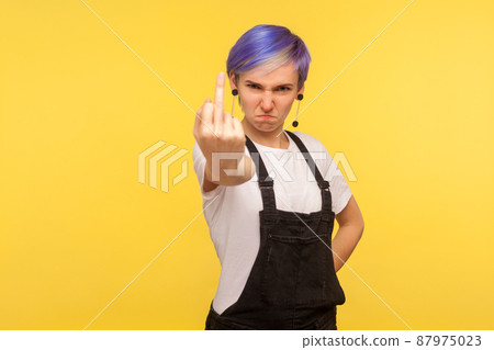Portrait of crazy angry hipster woman with violet hair in denim overalls showing middle finger, rude fuck off gesture, face full of hatred and aggression. isolated on yellow background, studio shot 87975023