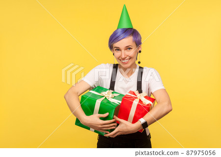 Portrait of happy joyful hipster girl with violet short hair wearing funny party cone and overalls embracing gift boxes, satisfied with presents, enjoying birthday party. yellow background studio shot Portrait of happy joyful hipster girl with violet short hair wearing funny party cone and overalls embracing gift boxes, satisfied with presents, enjoying birthday party. yellow background studio shot 87975056