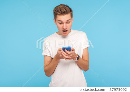 Portrait of amazed astonished man in white t-shirt using cellphone with surprised expression, shocked by message in social network, mobile communication. indoor studio shot isolated on blue background Portrait of amazed astonished man in white t-shirt using cellphone with surprised expression, shocked by message in social network, mobile communication. indoor studio shot isolated on blue background 87975092