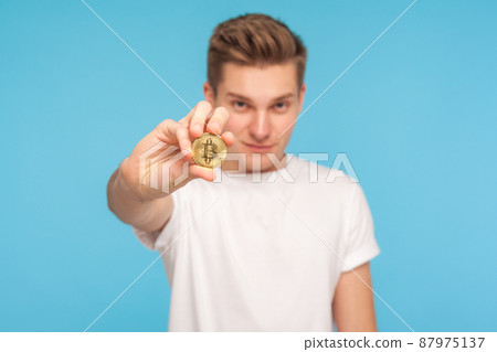 Portrait of young trendy rich man in white t-shirt holding golden bitcoin and looking at camera with clever expression, confident in cryptocurrency. indoor studio shot isolated on blue background Portrait of young trendy rich man in white t-shirt holding golden bitcoin and looking at camera with clever expression, confident in cryptocurrency. indoor studio shot isolated on blue background 87975137