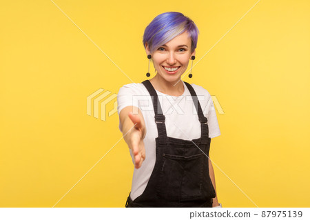 Nice to meet you. Portrait of cute positive hipster woman with violet short hair in denim overalls giving hand to handshake with friendly toothy smile. isolated on yellow background, studio shot 87975139