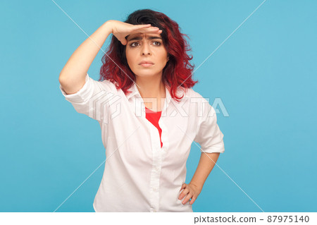 Distant future. Portrait of hipster woman with red hair holding hand over eyes and looking far away, expecting waiting something with worried expression. indoor studio shot isolated on blue background Distant future. Portrait of hipster woman with red hair holding hand over eyes and looking far away, expecting waiting something with worried expression. indoor studio shot isolated on blue background 87975140
