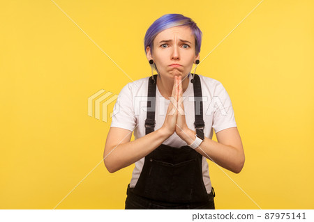 Portrait of upset hipster woman with violet short hair in denim overalls cupped hands in praying gesture, begging, apologizing, asking something with hopeful eyes. yellow background, studio shot 87975141