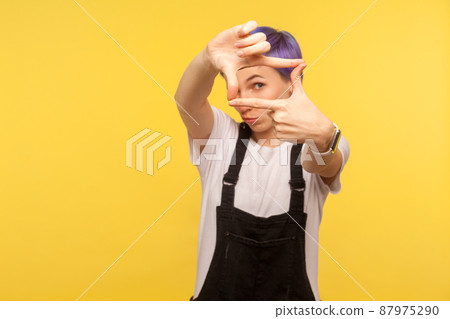 Portrait of nice focused hipster woman with violet short hair in denim overalls looking at camera through photo frame shaped with fingers, capturing moment. isolated on yellow background, studio shot 87975290