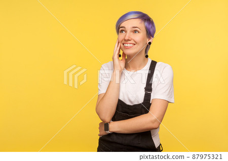 Portrait of happy beautiful hipster girl with violet short hair in denim overalls holding palm near face and dreaming pleasant fantasies, sweet memories. isolated on yellow background, studio shot Portrait of happy beautiful hipster girl with violet short hair in denim overalls holding palm near face and dreaming pleasant fantasies, sweet memories. isolated on yellow background, studio shot 87975321
