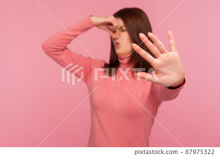 Confused brunette woman holding breath with fingers on nose showing stop gesture, dont come closer, shocked with disgusting stink. Indoor studio shot isolated on pink background Confused brunette woman holding breath with fingers on nose showing stop gesture, dont come closer, shocked with disgusting stink. Indoor studio shot isolated on pink background 87975322
