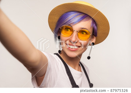 Portrait of beautiful glamour hipster girl with violet hair in hat and glasses taking selfie, looking at camera with toothy smile POV, point of view of photo. isolated on white background, studio shot 87975326