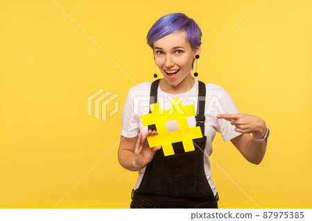 Hashtag, internet trends. Portrait of cool stylish hipster girl with violet short hair in denim overalls pointing finger at big hash sign, social network. isolated on yellow background, studio shot 87975385