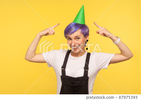 Birthday holiday. Portrait of positive stylish hipster girl with violet short hair in overalls pointing at funny party cone on her head and smiling at camera. isolated on yellow background studio shot 87975393