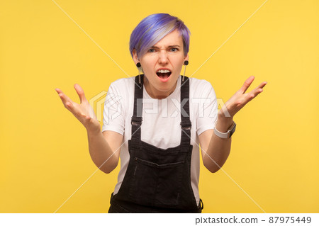 How could you. Portrait of outraged hipster woman with violet hair in overalls standing in bewilderment with raised hands, don't understand situation, indignant gesture. yellow background, studio shot How could you. Portrait of outraged hipster woman with violet hair in overalls standing in bewilderment with raised hands, don't understand situation, indignant gesture. yellow background, studio shot 87975449