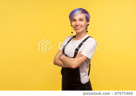 Portrait of positive cute hipster woman with violet dyed short hair in denim overalls smiling at camera and crossing hands, stylish fashionable appearance. isolated on yellow background, studio shot 87975460