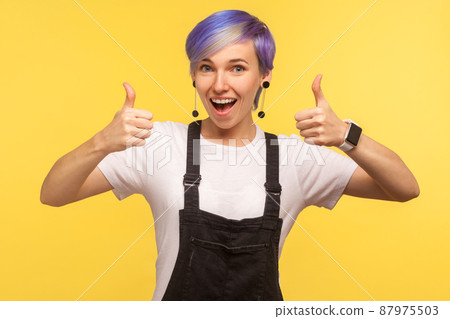 Like. Portrait of optimistic happy hipster woman with violet dyed short hair in denim overalls showing double thumbs up and saying wow yes, wide open mouth. isolated on yellow background, studio shot Like. Portrait of optimistic happy hipster woman with violet dyed short hair in denim overalls showing double thumbs up and saying wow yes, wide open mouth. isolated on yellow background, studio shot 87975503