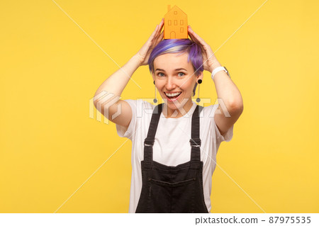 Portrait of nice fashionable hipster girl with violet short hair in denim overalls holding orange paper house on her head and smiling genuinely at camera. studio shot isolated on yellow background 87975535