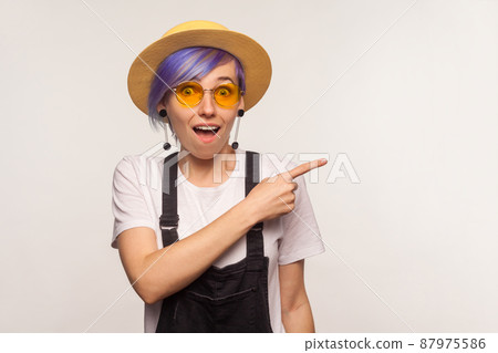 Look there. Portrait of surprised fashionable hipster girl with violet hair in sunglasses and hat pointing to the side, copy space for promotional text, advertisement. white background, studio shot 87975586
