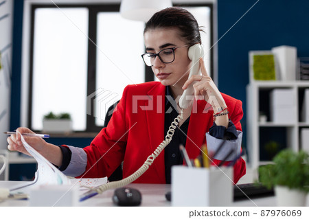 Businesswoman with glasses looking at papers in phonecall. Entrepreneur in red jacket sitting at desk analyzing clipboard with chart. Small business owner in startup office in phone conversation. 87976069