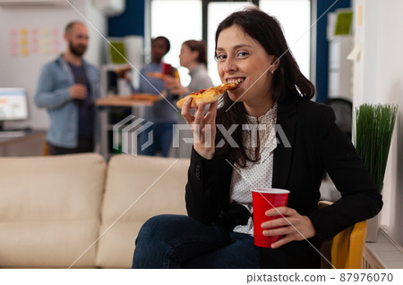 Portrait of woman eating pizza after work hours at party celebration in business office. Cheerul worker feeling relaxed at meeting with drinks, food, fun and leisure activity. Portrait of woman eating pizza after work hours at party celebration in business office. Cheerul worker feeling relaxed at meeting with drinks, food, fun and leisure activity. 87976070