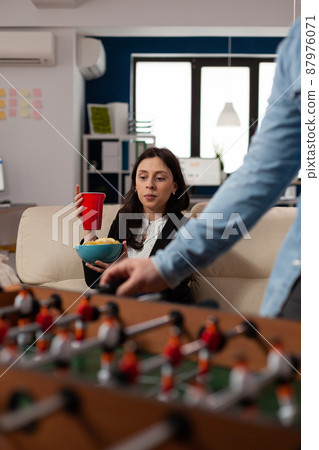 Portrait of young woman holding glass of beer at after work celebration, looking at foosball game play. Cheerful person enjoying drinks part after hours, celebrating with office workmates. 87976071