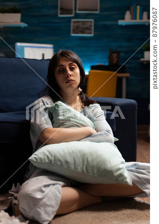 Portrait of depressed woman with mental health problems sitting on floor and hugging pillow in living room. Anxious sad person dealing with chronic disease difficulties and depression at home. 87976087