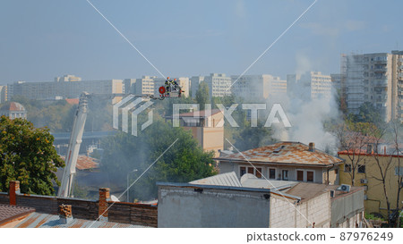 Firefighters using platform truck to help with house on fire. Firemen from fire department trying to put out flames and smoke from burning building in neighbourhood. Men extinguishing fumes Firefighters using platform truck to help with house on fire. Firemen from fire department trying to put out flames and smoke from burning building in neighbourhood. Men extinguishing fumes 87976249