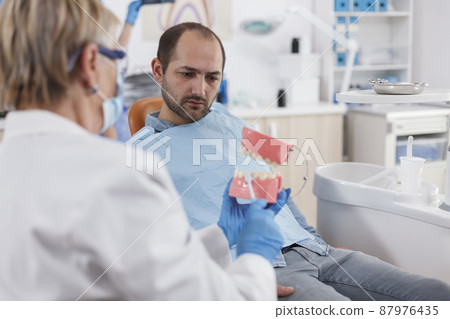 Specialist stomatologist doctor with face mask holding jaw ceramic model explaining oral hygiene to man patient during dentistry examination in stomatological office room. Stomatology concept 87976435