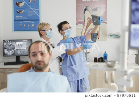 Portrait of patient with toothache waiting for medical procedure during stomatological consultation in dentistry office room. In background medical team analyzing teeth radiography. Medicine service 87976611