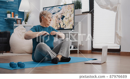 Senior woman following training video program on laptop to do workout and physical activity. Retired person watching online lesson with trainer to exercise fitness and gymnastics on mat. 87976689